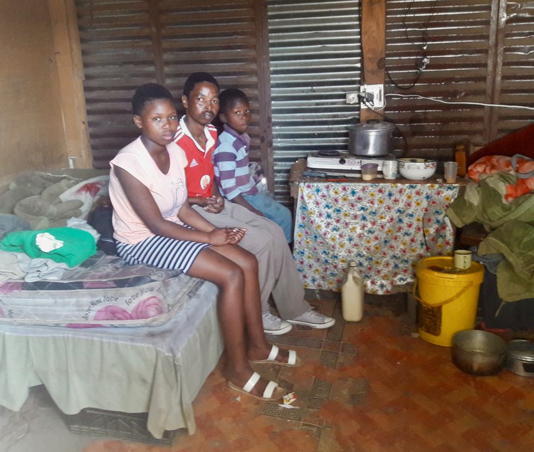 Photo of three people sitting on a bed in a shack