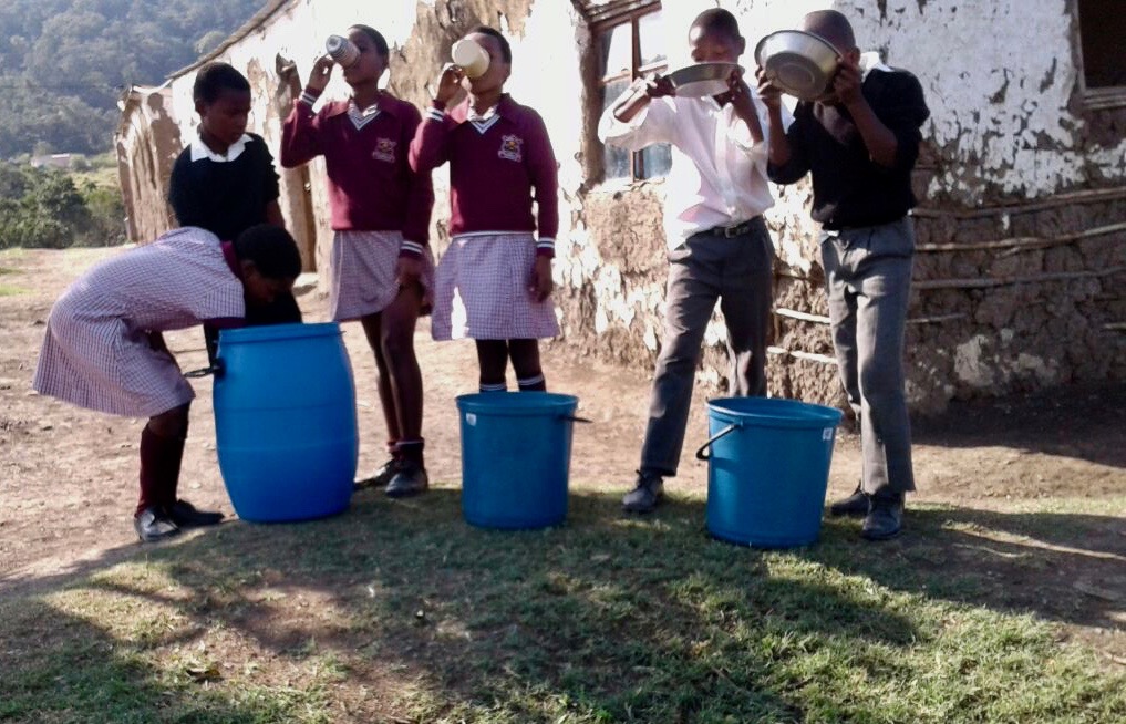 Photo of school children drinking water from blue plastic drums